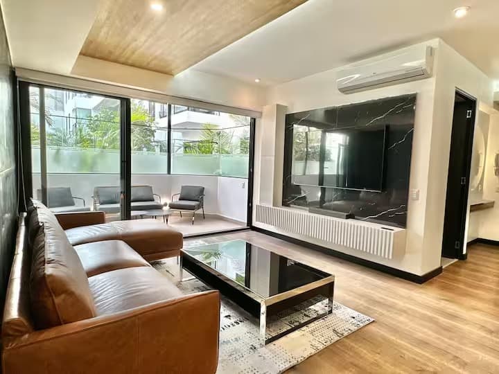 Sala de un departamento con techo de madera, sofá de cuero y terraza al parque